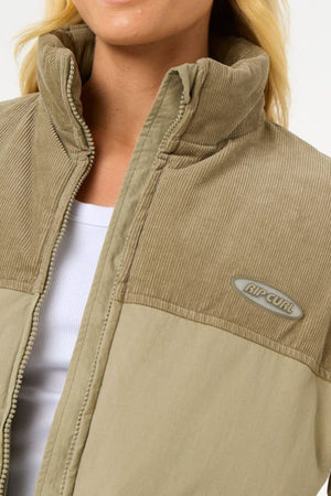 Close-up of a person wearing a light brown Rip Curl High Tide Mixed Cord Puffer, featuring corduroy texture on the top half, smooth fabric below, and an embroidered logo. A white shirt is visible underneath.