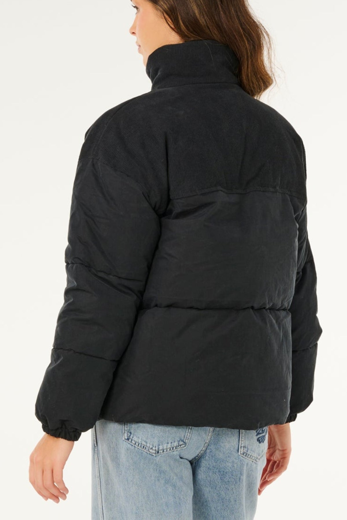 A person with long brown hair is seen from behind, wearing the Rip Curl High Tide Mixed Cord Puffer with a high collar and light blue jeans. The background is plain white.
