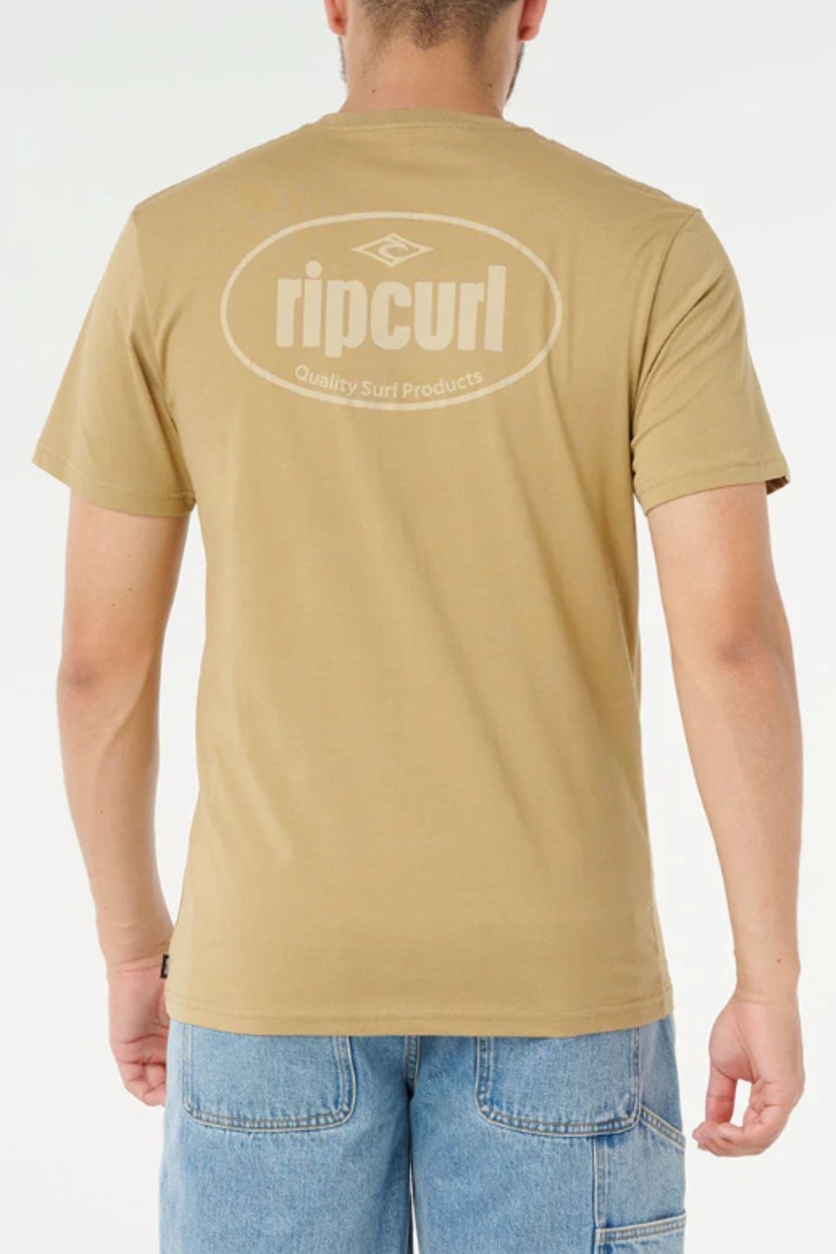 A person is seen from the back wearing a tan Rip Curl Everyday Tee with "Rip Curl" and "Quality Surf Products" in white on the upper back, paired with light blue denim shorts.