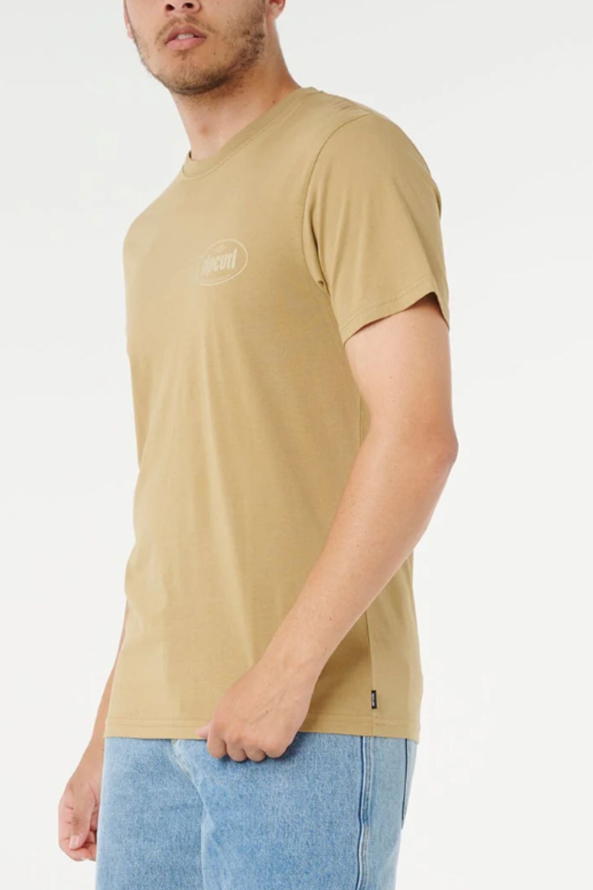 Wearing the Rip Curl Everyday Tee in tan—a classic short-sleeve cotton t-shirt with a small chest logo—a person pairs it with light blue jeans and stands against a white background, their face partially out of frame.