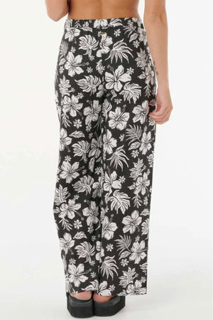 A person stands facing away in Rip Curl Keepsakes Beach Pant, featuring a high waist and bold white tropical floral print. The loose, breathable fabric pairs with black shoes, set against a plain, light background.