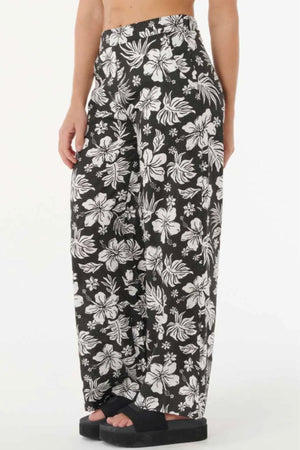Someone wearing the Rip Curl Keepsakes Beach Pant—loose-fitting, high-waist black pants with a white hibiscus and tropical floral print—paired with black platform sandals, stands against a plain white background.