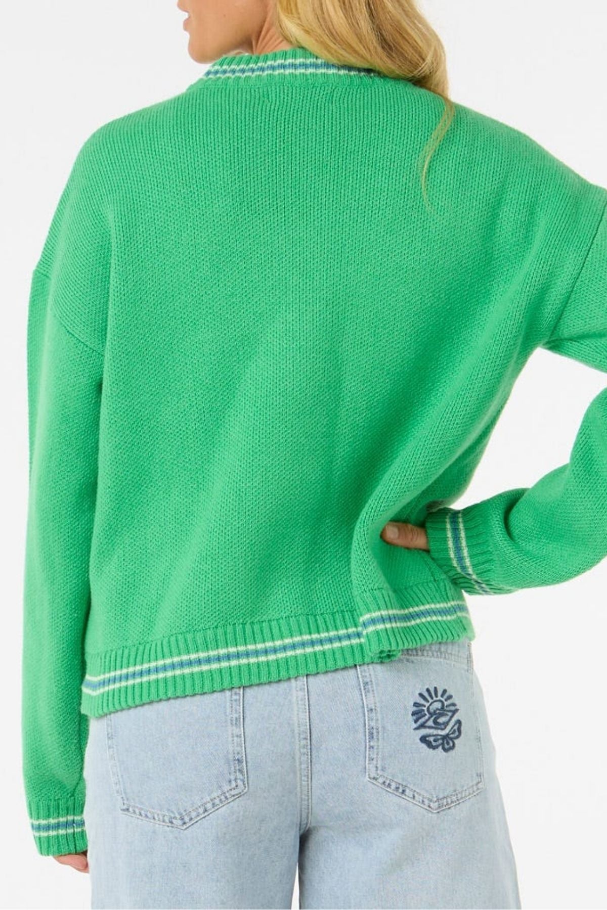 A person with long blonde hair stands with one hand on their hip, wearing a cozy, stylish bright green Rip Curl Las Dalias Sweater with white trim and light blue jeans featuring a blue sun and flower design on the back pocket.
