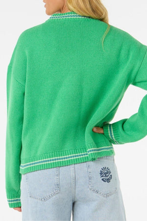 A person with long blonde hair stands with one hand on their hip, wearing a cozy, stylish bright green Rip Curl Las Dalias Sweater with white trim and light blue jeans featuring a blue sun and flower design on the back pocket.