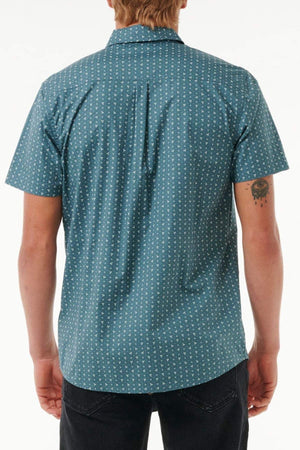A person with a tattoo on their left arm stands with their back to the camera, wearing black pants and a teal Rip Curl Micro Short Sleeve Shirt featuring a small white pattern.
