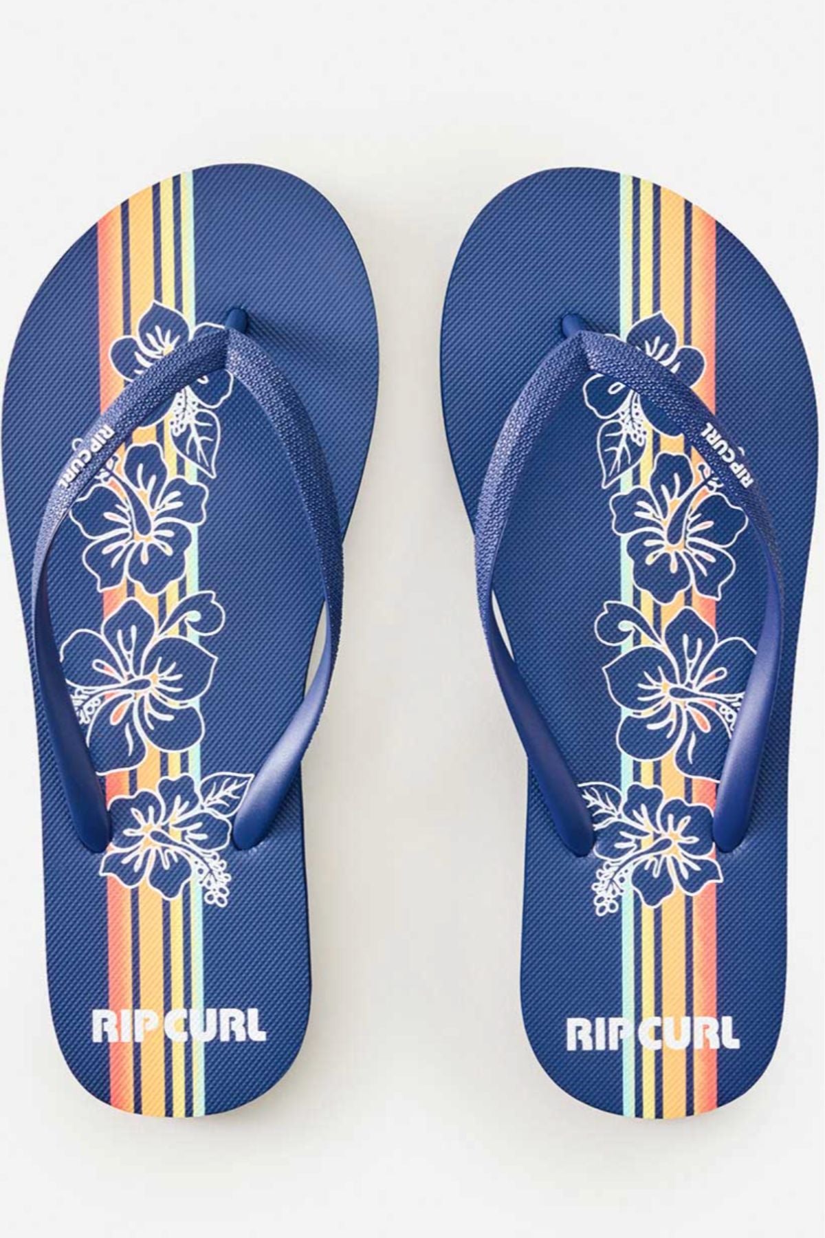 The Rip Curl Mixed Bloom Thongs feature a blue base with white hibiscus prints and bold yellow, orange, and red stripes on eco-friendly algae-blended EVA foam soles.