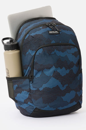 Rip Curl Ozone 30L Camo Check backpack featuring a dark topographic mountain pattern, with a laptop in the main compartment and a water bottle in the side mesh pocket.