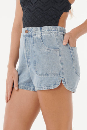 Woman wearing light blue Rip Curl Pacific Rider Denim Short with a curved hem and front pockets, paired with a sleeveless black textured top. One hand is in her pocket while the other rests by her side.