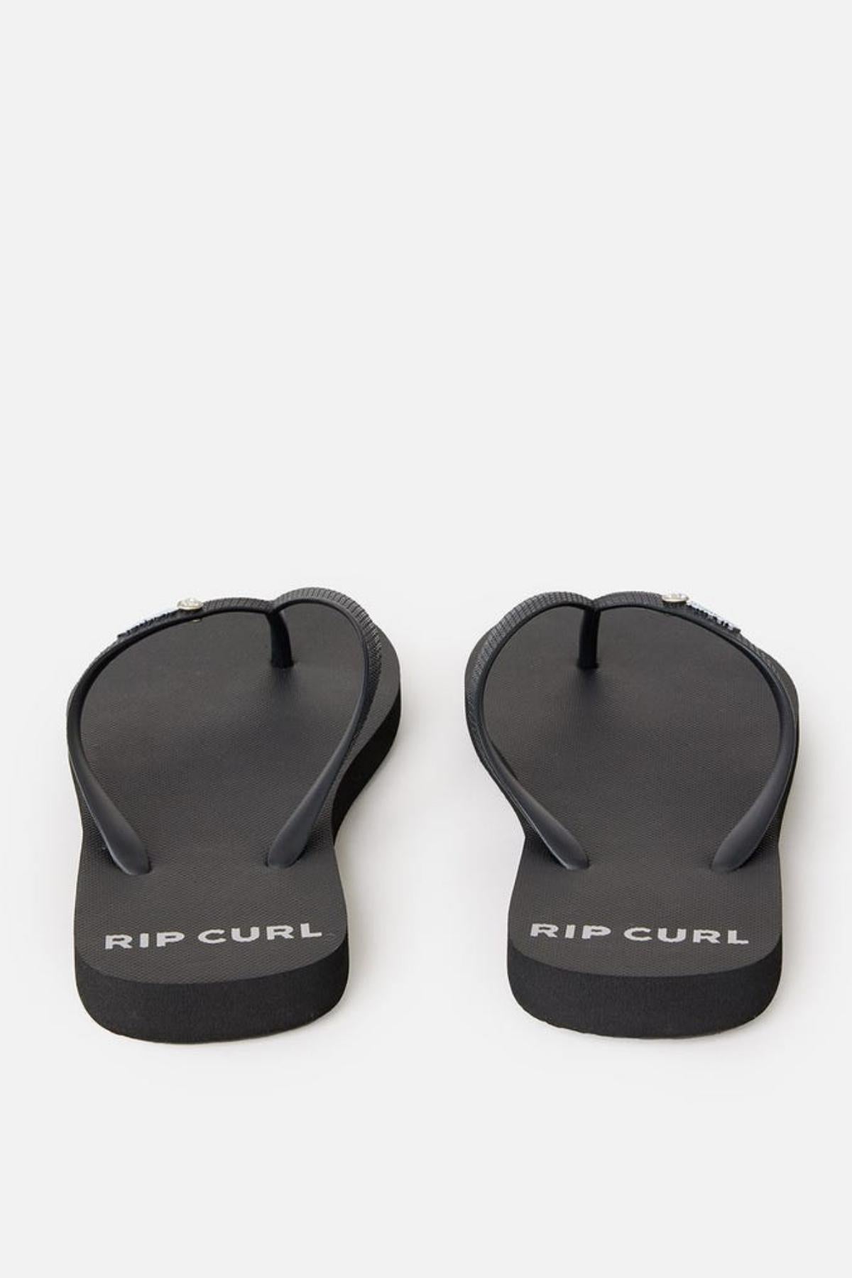 A pair of Rip Curl Premium Surf Bloom Thongs with the words "RIP CURL" printed in white on the footbeds. These black flip-flops feature a simple design with black straps and rest on a plain white background.