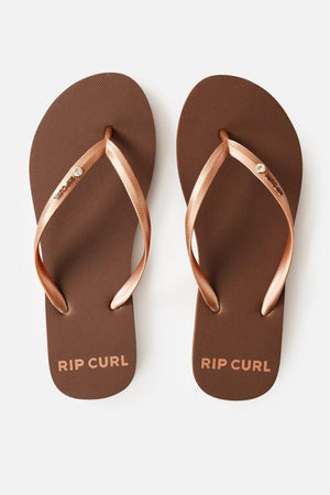 The Rip Curl Premium Surf Bloom Thongs feature a pair of brown flip-flops with gold straps. The text "RIP CURL" is elegantly printed in gold lettering on the footbed near the toe section of each sandal. Additionally, one of the straps on each sandal is adorned with a small circular metallic emblem. The background is white.
