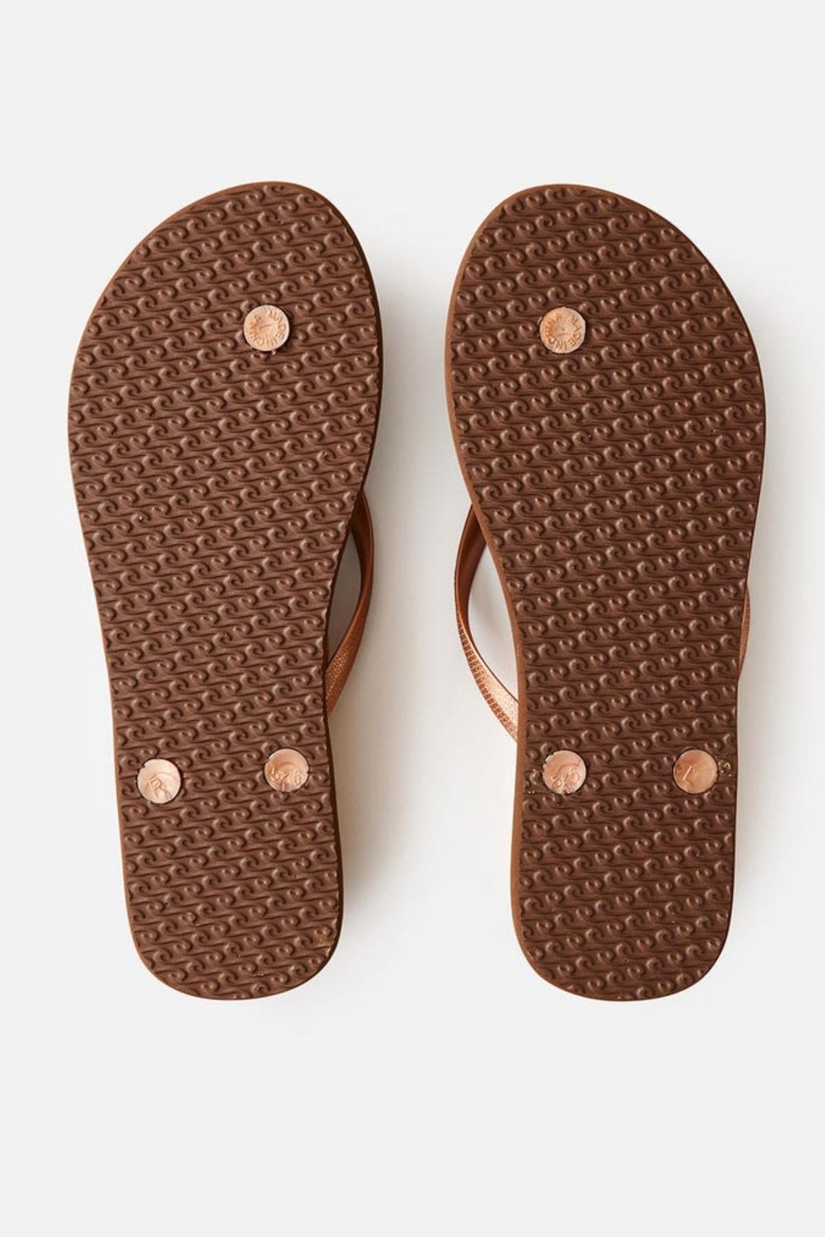The image shows the bottoms of a pair of Rip Curl Premium Surf Bloom Thongs with textured soles. The soles feature a pattern and small round rivets where the straps connect to the base. The thongs are placed side by side on a plain, white background.