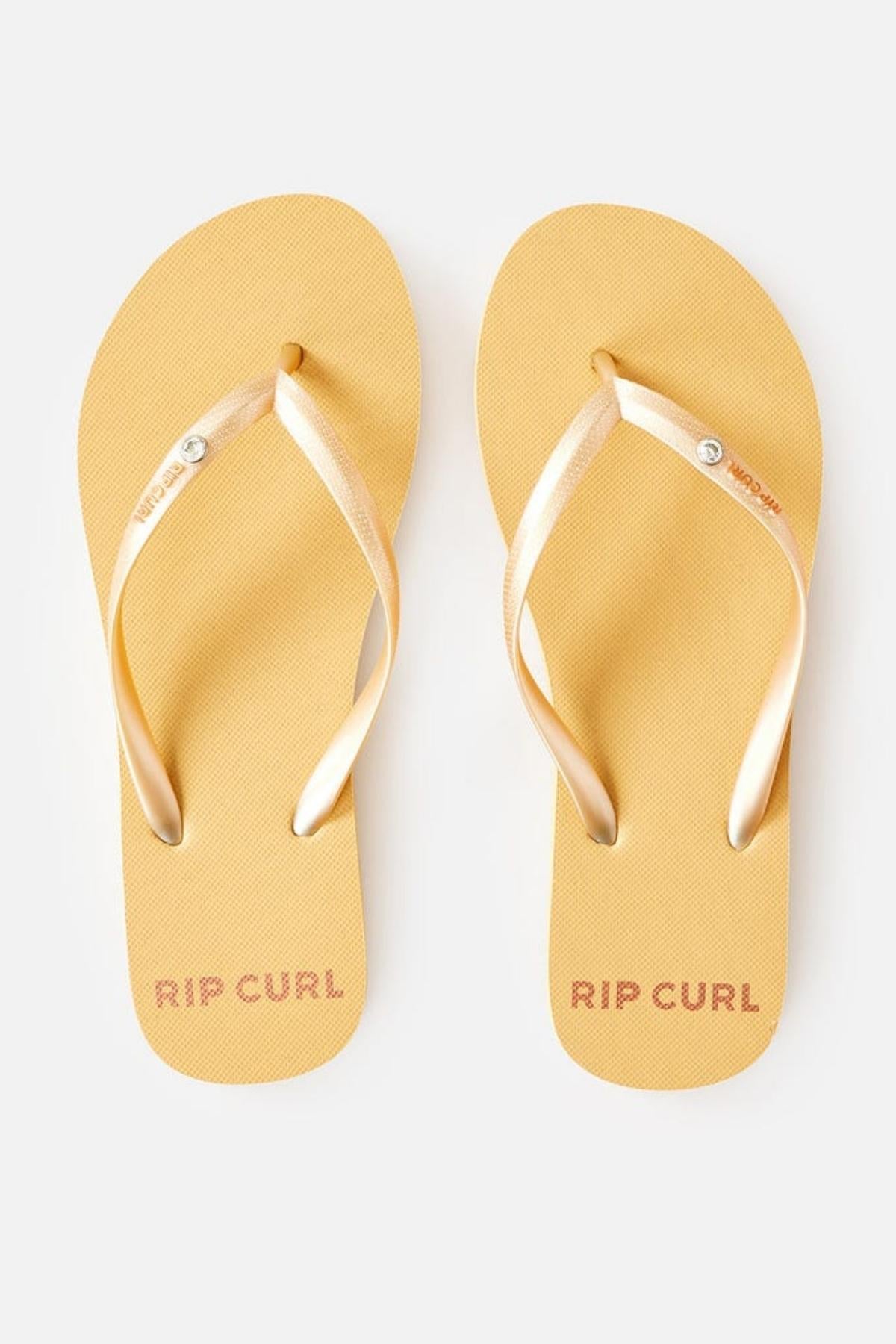 A pair of yellow Rip Curl Premium Surf Bloom Thongs with metallic gold straps is viewed from above. The right thong's strap features the Rip Curl logo. "RIP CURL" is printed in pink on the heel section of both thongs. The thongs are set against a plain white background.