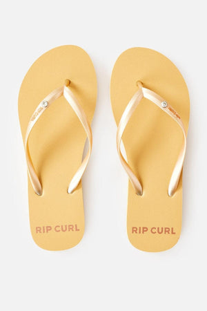 A pair of yellow Rip Curl Premium Surf Bloom Thongs with metallic gold straps is viewed from above. The right thong's strap features the Rip Curl logo. "RIP CURL" is printed in pink on the heel section of both thongs. The thongs are set against a plain white background.