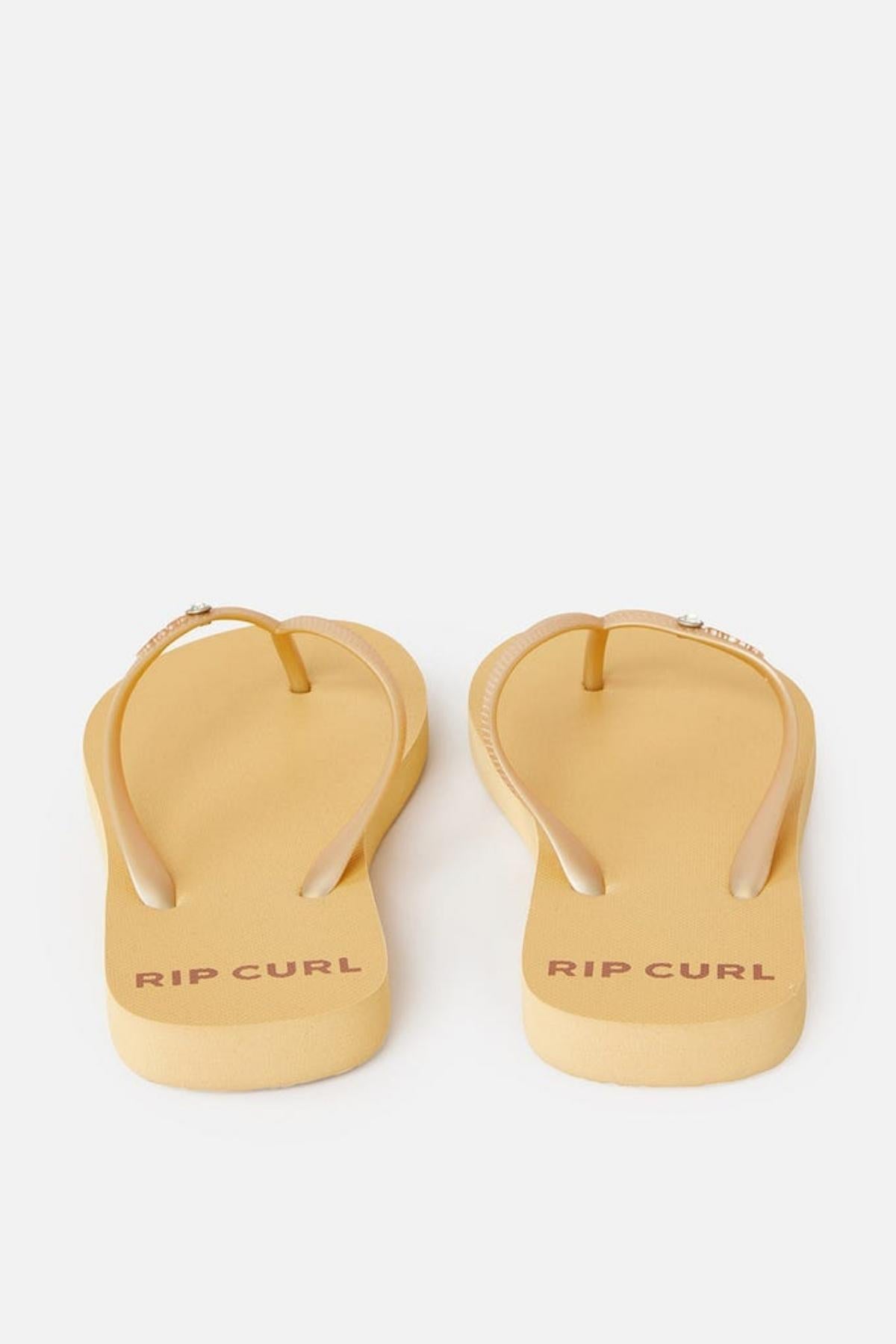 A pair of Rip Curl Premium Surf Bloom Thongs in beige with straps and the brand name "RIP CURL" printed in red on the footbeds. The thongs are positioned with the toe end facing away, showcasing the soles and strap design, set against a plain white background.
