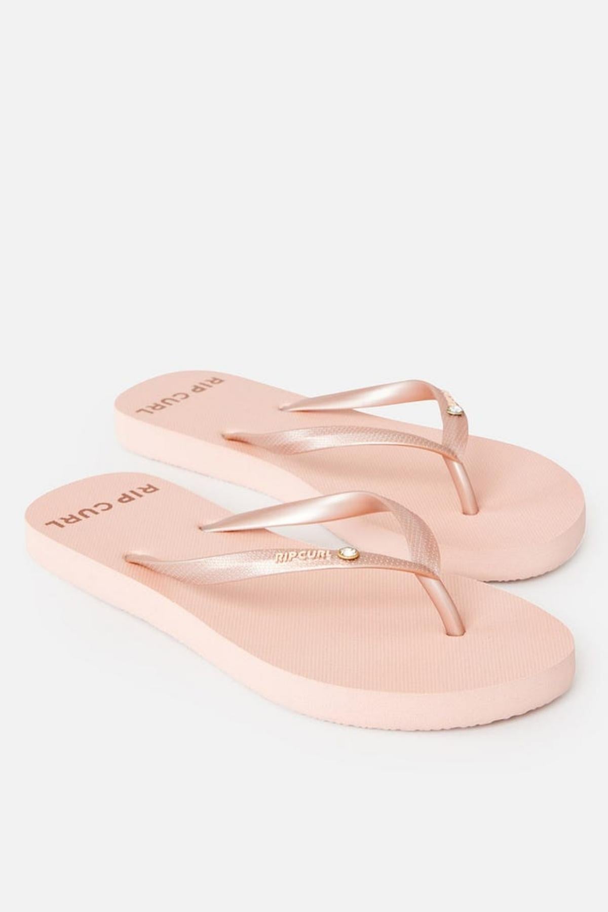 The Rip Curl Premium Surf Bloom Thongs are light pink flip-flops featuring a simple design. Both the footbeds and straps are pink, with the straps showcasing a small metallic detail. The "Rip Curl" branding is visible on the footbeds near the heels, all set against a white background.