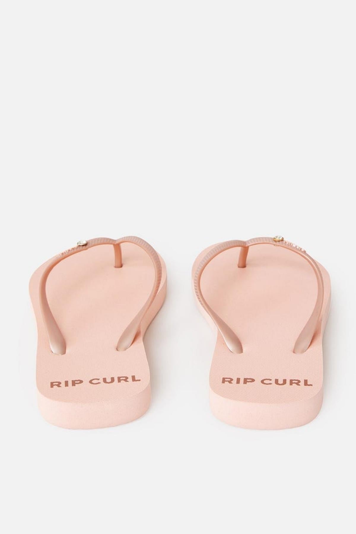 A pair of Rip Curl Premium Surf Bloom Thongs in light pink, featuring thin straps and "RIP CURL" printed on the heel of each sole, are displayed against a plain white background with the toe ends pointing towards the top of the image.