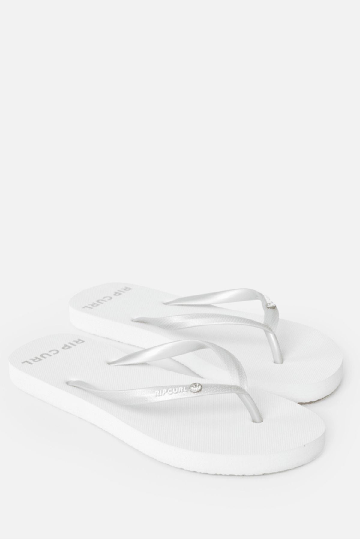 A pair of white Rip Curl Premium Surf Bloom Thongs with thin silver straps is displayed on a white background. Eco-friendly and ideal for the beach, they feature the "Rip Curl" logo printed on the footbed near the heel.