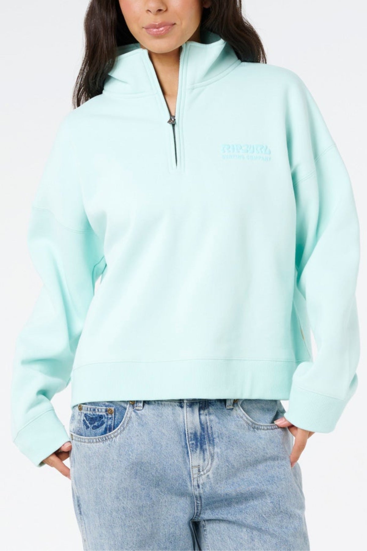 Wearing the Rip Curl Surf Puff 1/4 Zip pullover in light blue with long sleeves and faded blue jeans, this person stands with one hand in their pocket against a plain white background for an easy surf style.