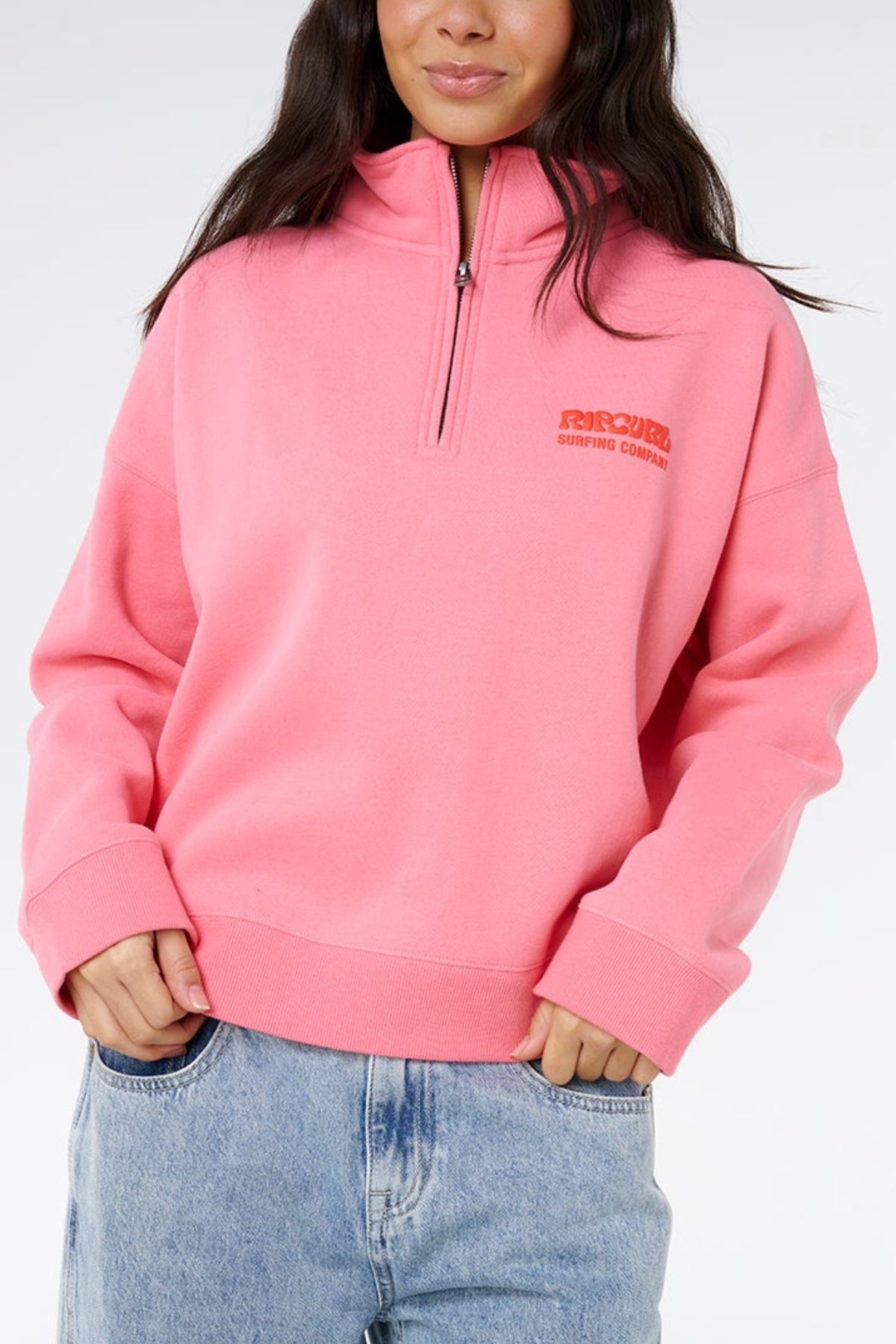 A woman models the Rip Curl Surf Puff 1/4 Zip pullover in bright pink, featuring the "Rip Curl Surfing Company" chest logo, paired with light blue jeans against a plain background for a laid-back surf-inspired look.