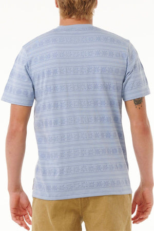 A person with light hair is shown from the back, wearing the Rip Curl Saltwater Culture Stripey Tee in light blue with jacquard stripes and tan corduroy pants. The 100% cotton shirt reveals a tattoo on their upper left arm.
