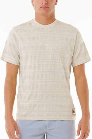 A man wearing a Rip Curl Saltwater Culture Stripey Tee with subtle horizontal designs and light blue pants stands against a plain white background.