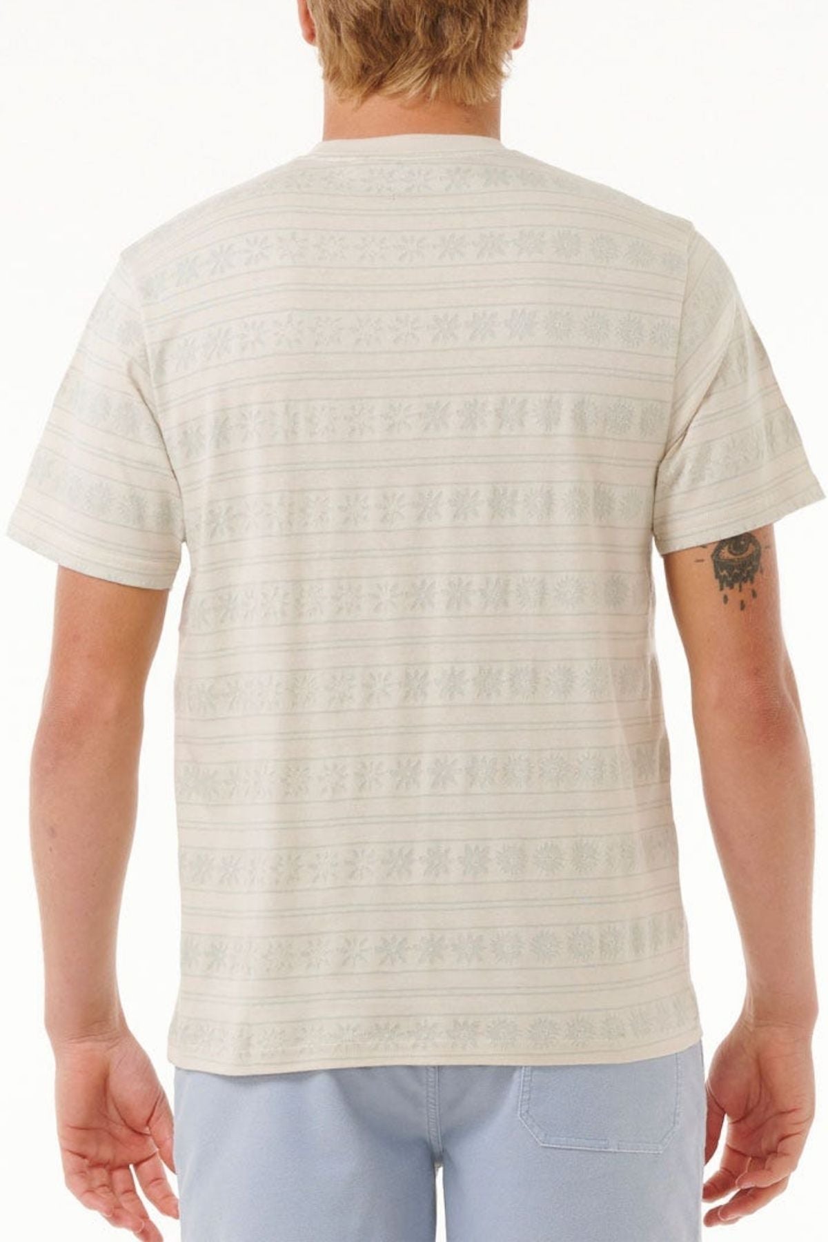 Viewed from the back, a person with short blonde hair wears the Rip Curl Saltwater Culture Stripey Tee paired with light blue shorts. A small tattoo is visible on their left arm against a plain white backdrop.
