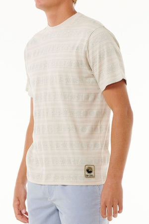 A person dressed in a Rip Curl Saltwater Culture Stripey Tee with light blue pants stands against a plain background. The tee has a small design patch near the hem, capturing the Saltwater Culture vibe. The person's face is not visible.