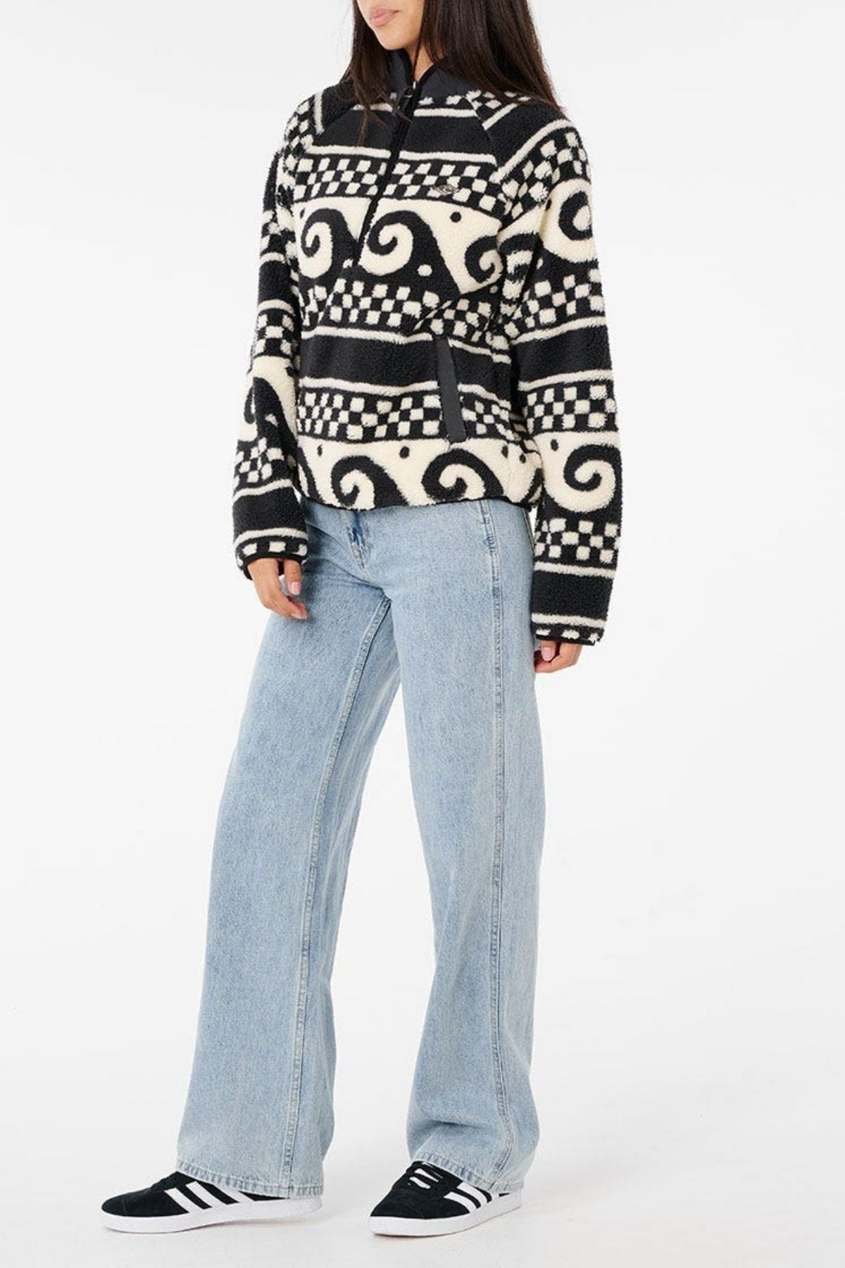 Someone wearing a Rip Curl San Carlos Polar Fleece in black and white, paired with light blue wide-leg jeans and black sneakers with white stripes, stands against a plain white background.