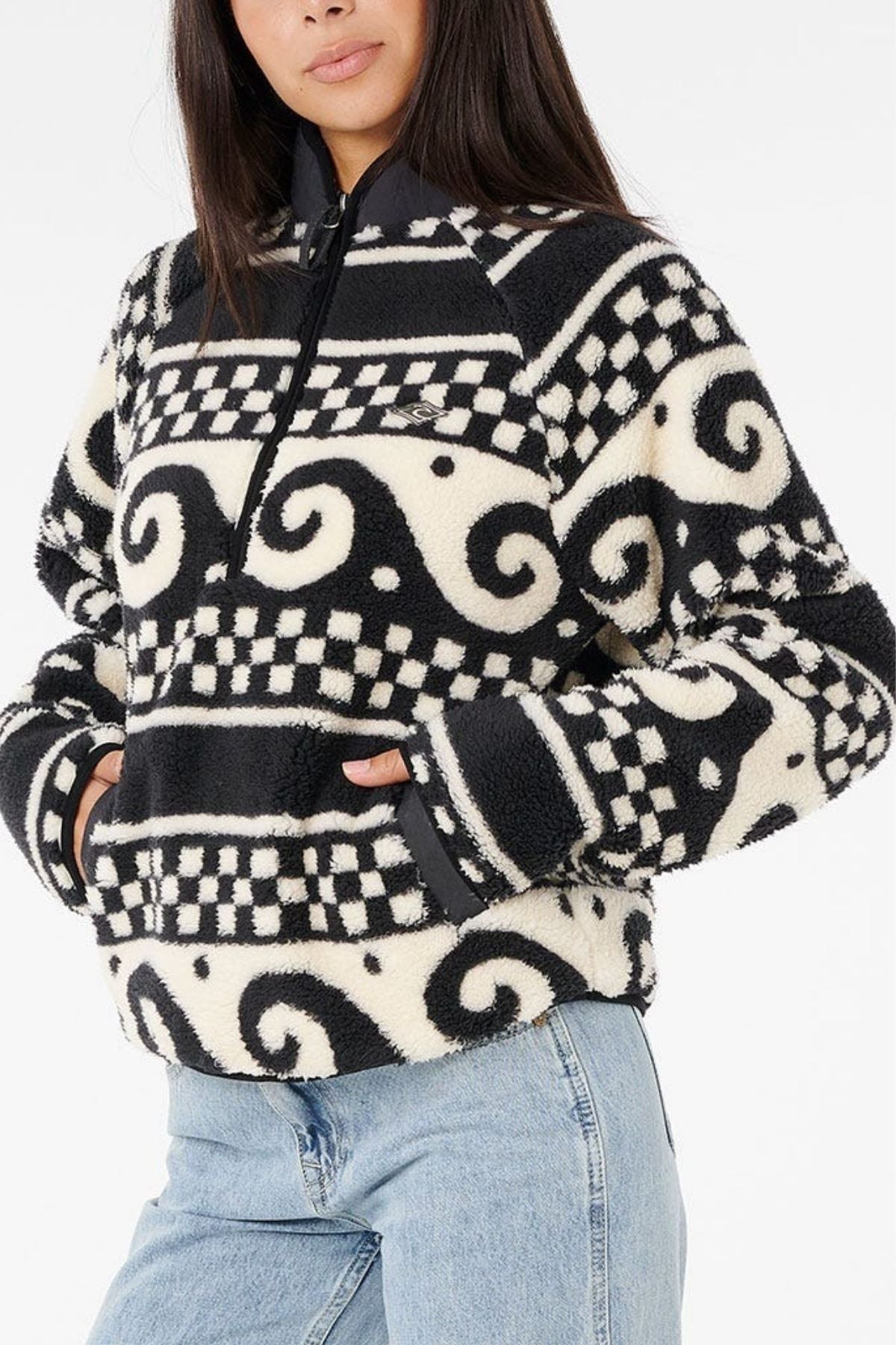 A woman with long dark hair wears the Rip Curl San Carlos Polar Fleece, featuring black and white swirls and checkerboard patterns, paired with light blue jeans. Her hands are tucked into the pullover's pockets.
