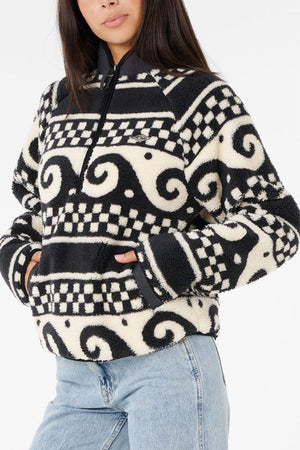 A woman with long dark hair wears the Rip Curl San Carlos Polar Fleece, featuring black and white swirls and checkerboard patterns, paired with light blue jeans. Her hands are tucked into the pullover's pockets.