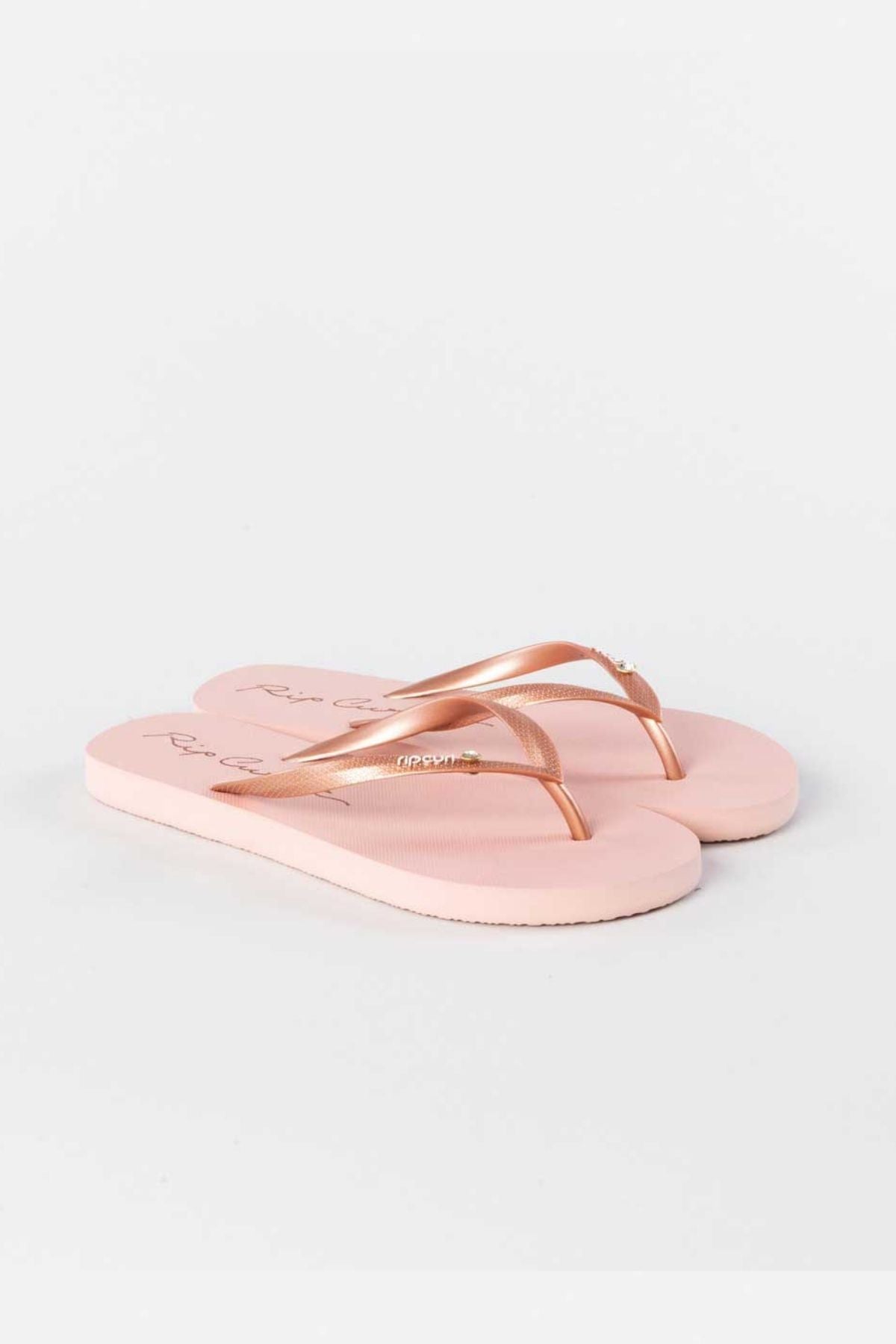 A pair of Rip Curl Script Wave Bloom Thongs in light pink, crafted from eco-friendly materials with slim straps and a slight sheen, showcased against a white background.