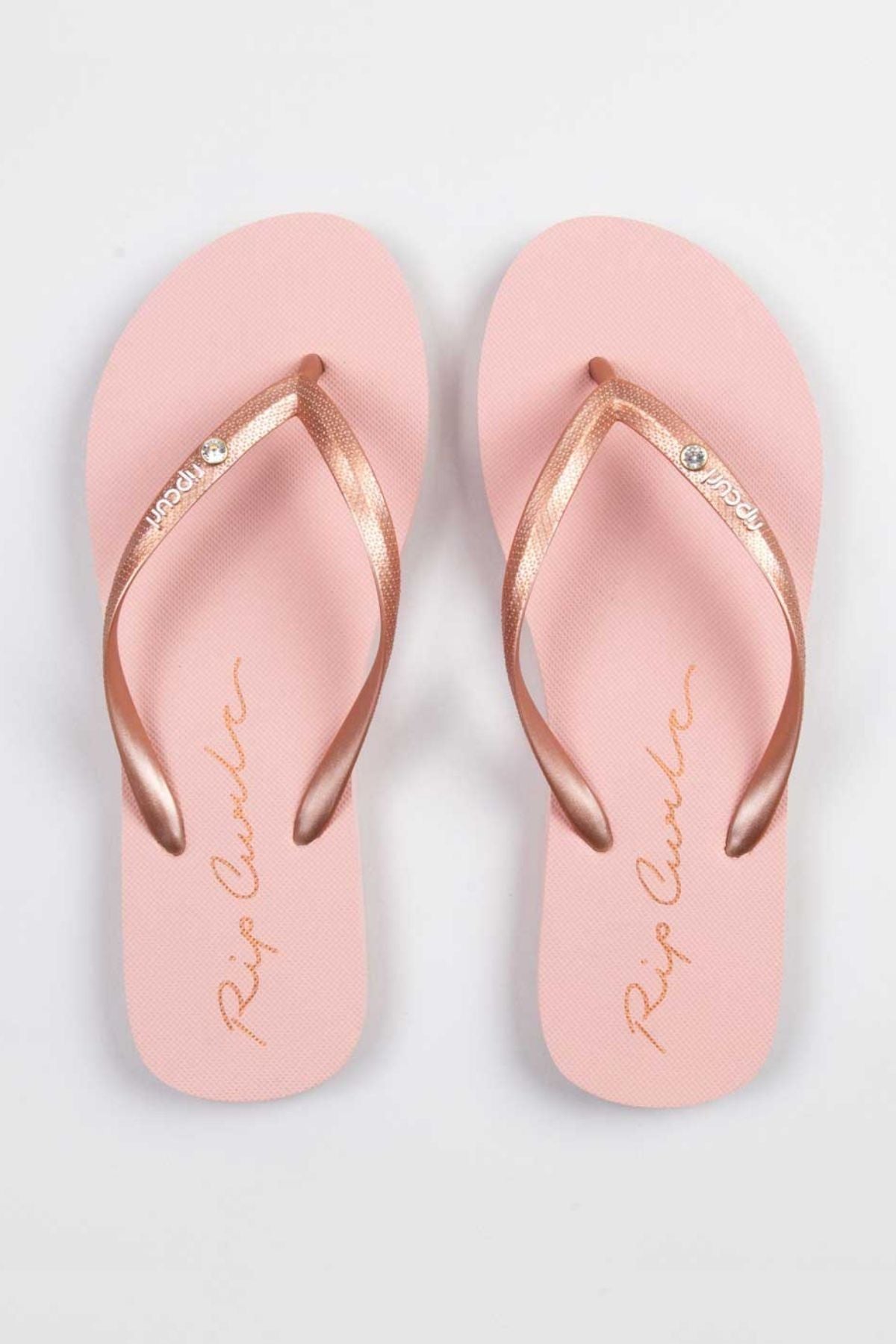 The Rip Curl Script Wave Bloom Thongs feature a pair of pink sandals with metallic rose gold straps. The EVA footbeds showcase a cursive logo design, accompanied by the brand name "Rip Curl" in gold, made from eco-friendly materials. They are elegantly displayed on a light gray background.