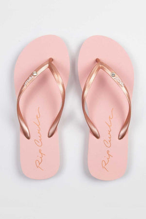 The Rip Curl Script Wave Bloom Thongs feature a pair of pink sandals with metallic rose gold straps. The EVA footbeds showcase a cursive logo design, accompanied by the brand name "Rip Curl" in gold, made from eco-friendly materials. They are elegantly displayed on a light gray background.