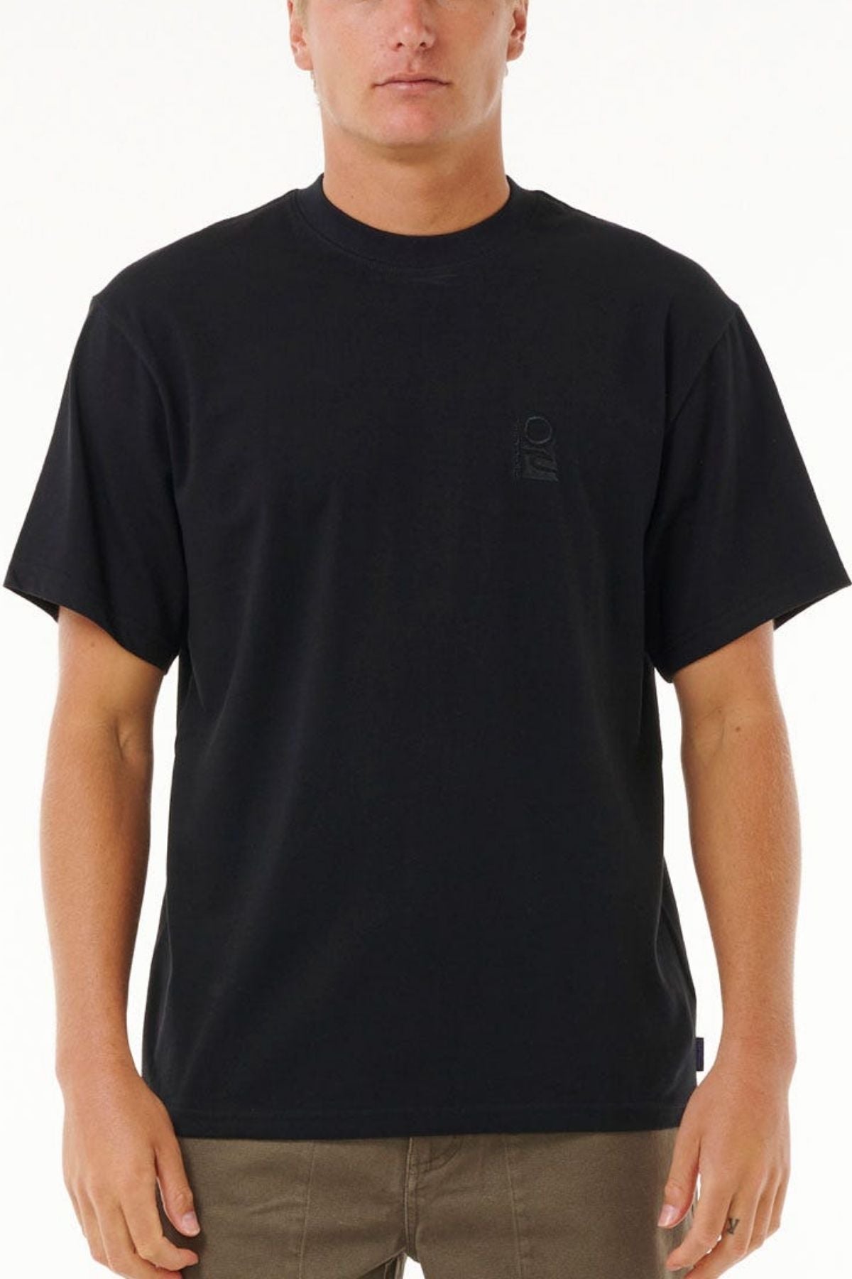 A person in a plain black Rip Curl Search Embroidery Tee made from 100% cotton and brown pants stands facing forward against a white background. The tee features a subtle, barely visible chest design.