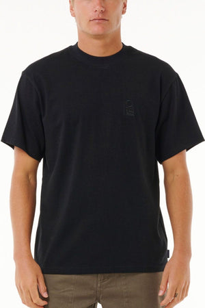 A person in a plain black Rip Curl Search Embroidery Tee made from 100% cotton and brown pants stands facing forward against a white background. The tee features a subtle, barely visible chest design.