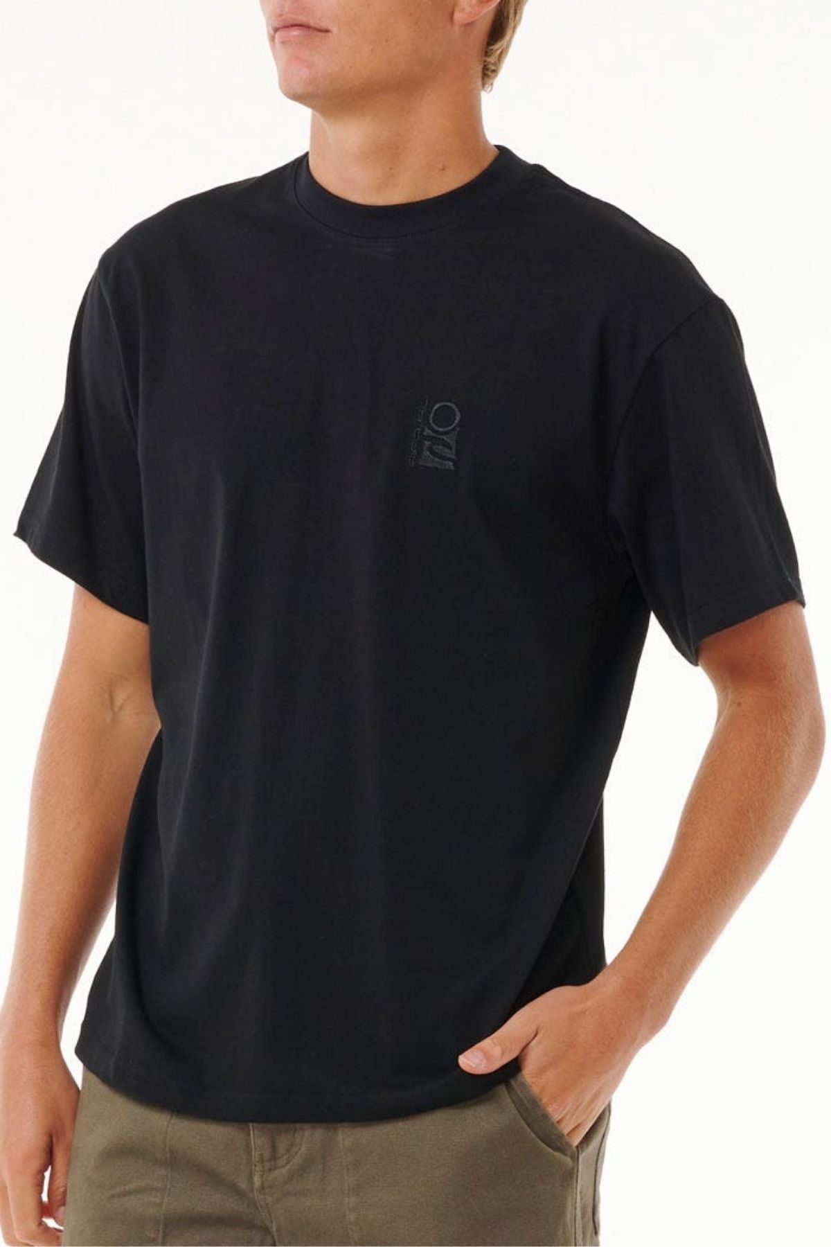 A person wearing a loose-fitting Rip Curl Search Embroidery Tee in black, made from 100% cotton with a small chest logo, and olive green pants stands with one hand in their pocket against a plain white background.