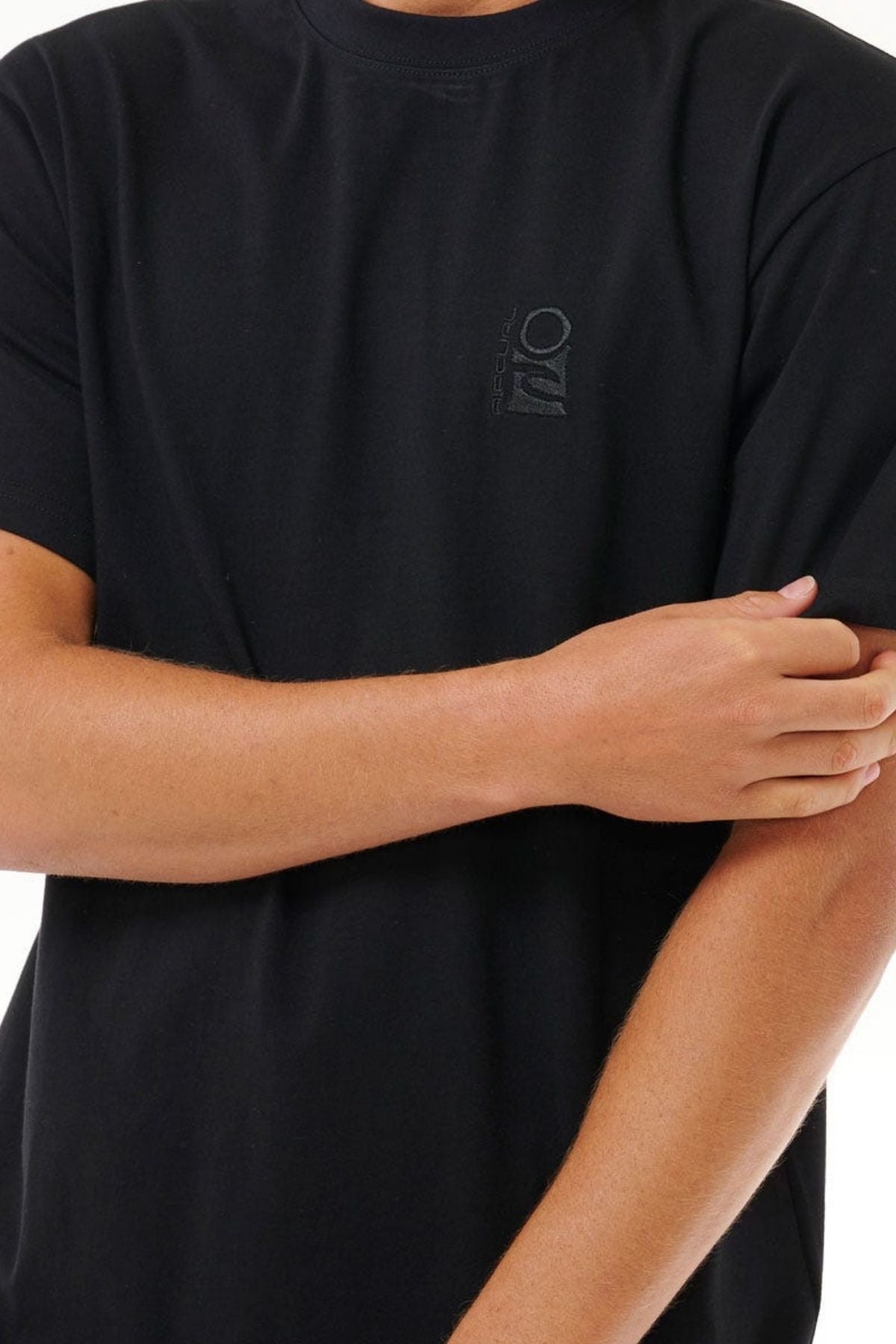 A person wearing a plain black Rip Curl Search Embroidery Tee, made from 100% cotton with a small chest logo, stands against a white background with arms crossed at the elbow and head not visible.