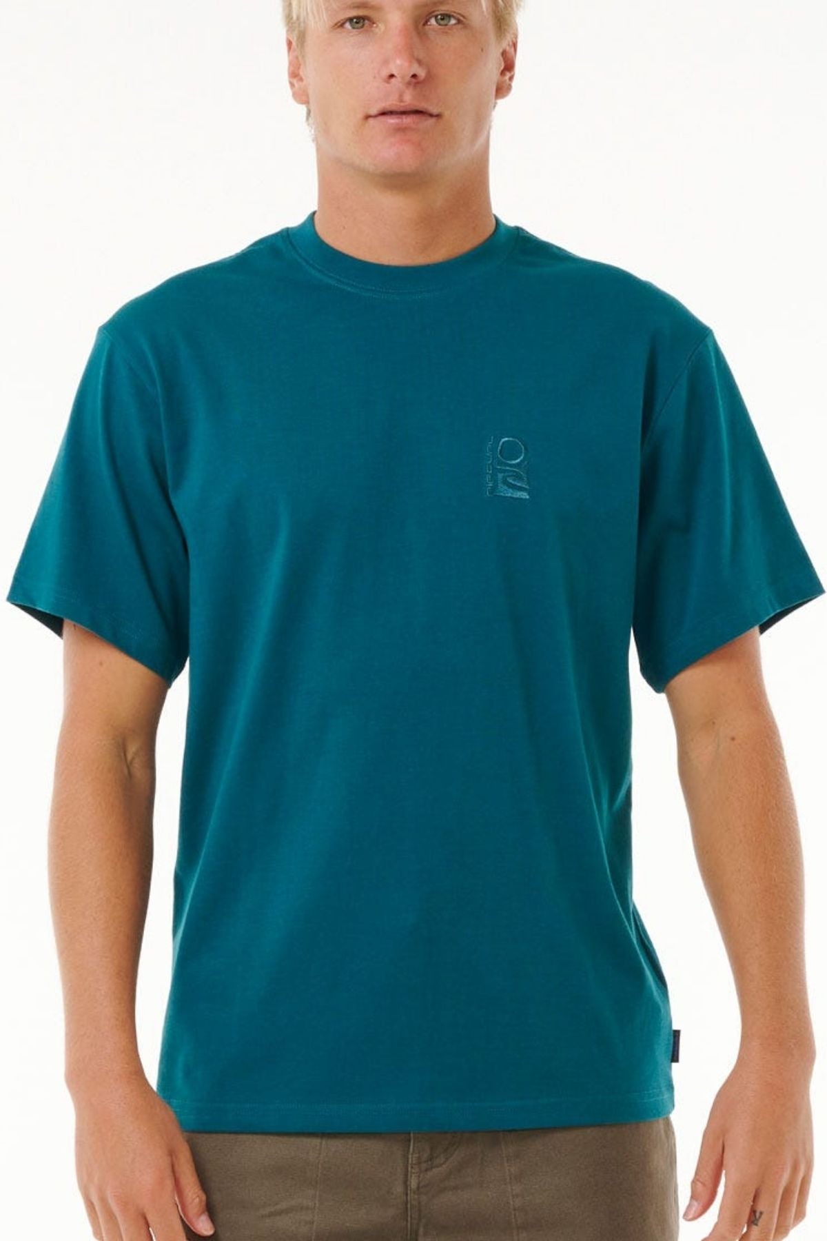 A person wearing a teal Rip Curl Search Embroidery Tee with a small logo on the left chest stands facing forward, sporting short blonde hair and brown pants against a plain white background.