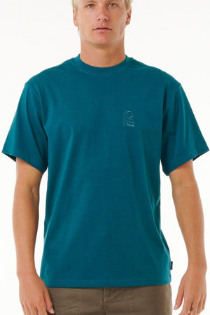 A person wearing a teal Rip Curl Search Embroidery Tee with a small logo on the left chest stands facing forward, sporting short blonde hair and brown pants against a plain white background.
