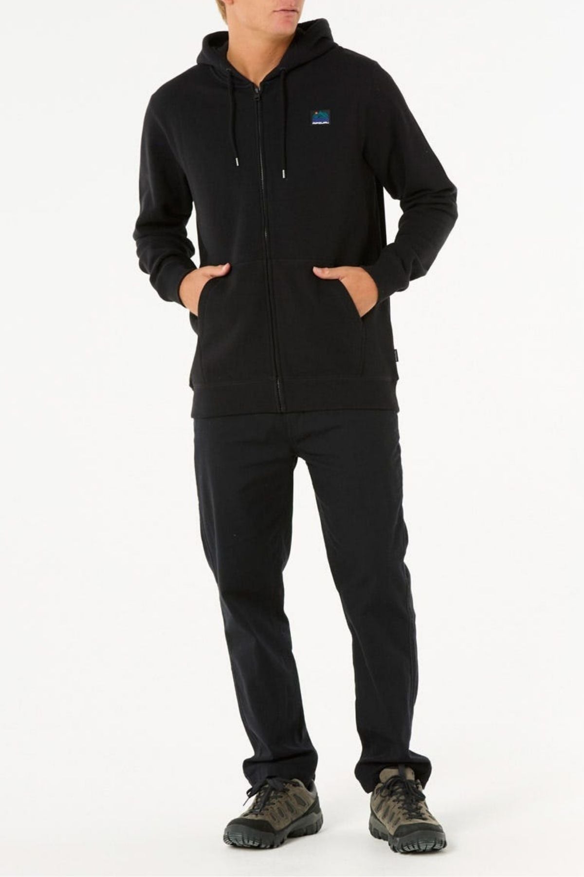 A person stands with hands in the pockets of the Rip Curl Search Zip Thru Hood, wearing dark pants and black shoes against a plain white background. Made from a soft cotton polyester blend. Their face is not visible.