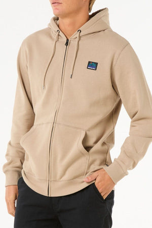 A person wears a tan Rip Curl Search Zip Thru Hood with a small chest patch and metal zipper, paired with black pants, standing against a plain white background with one hand in their pocket.