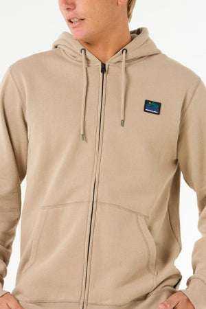 A person wearing the Rip Curl Search Zip Thru Hood in light beige, featuring a small mountain patch on the left chest, metal zipper, drawstrings, and front pockets, is facing slightly to the side.