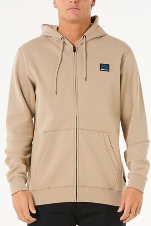 A person wearing a beige Rip Curl Search Zip Thru Hood featuring a small chest patch, hood with drawstrings, front pockets, metal zipper closure, and black pants stands against a plain white background.
