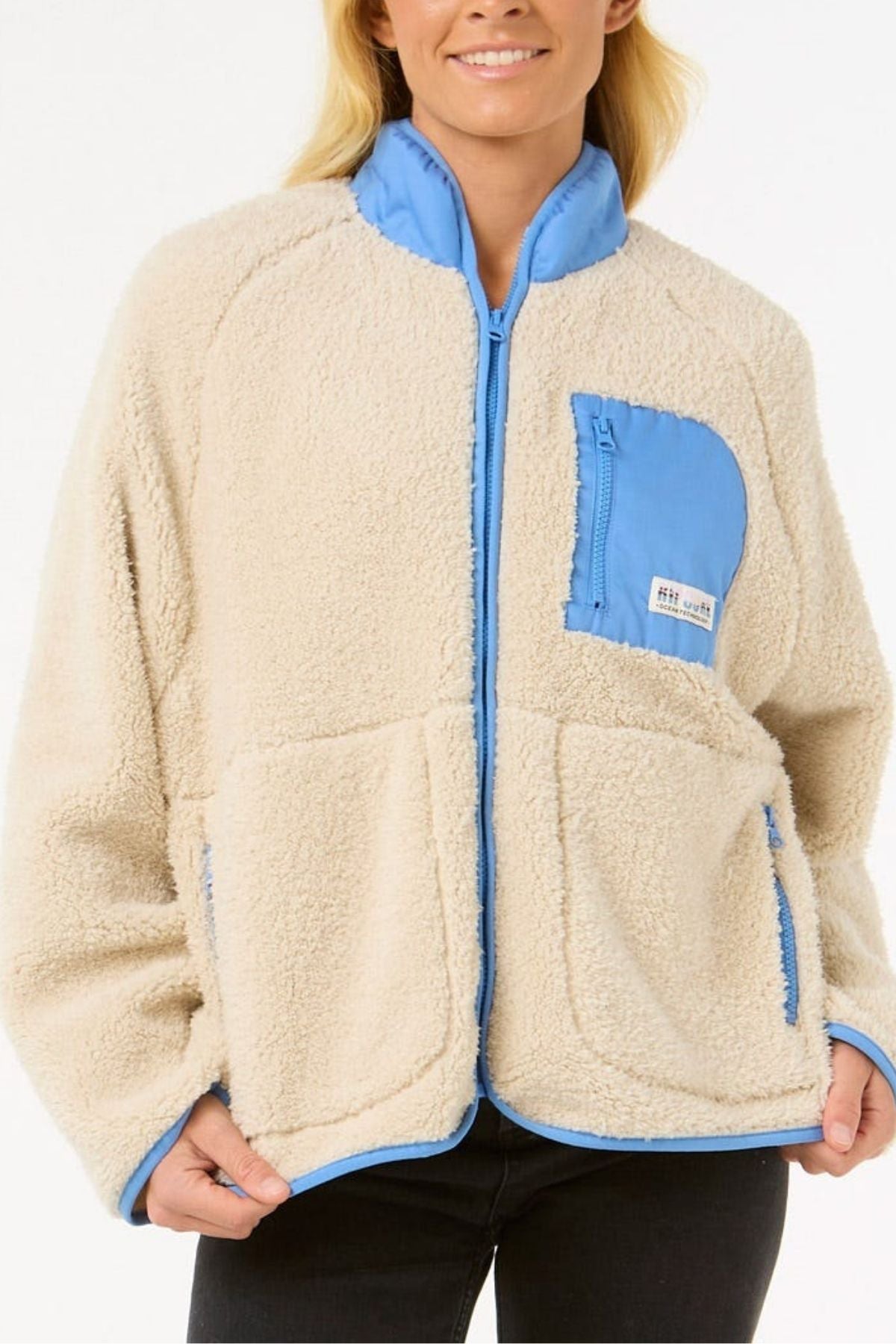 A person wears the Rip Curl Shore Break Polar Fleece—a beige, relaxed-fit pullover with light blue trim, blue zipper, and chest pocket—made from 100% Polyester 300GSM microfleece. Only their lower face and upper body are visible against a white background.