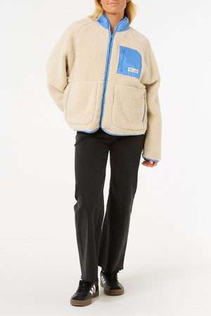 Someone stands against a plain white background wearing the Rip Curl Shore Break Polar Fleece, a cream-colored sherpa jacket with light blue details, paired with black pants and black sneakers.
