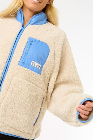 A person wearing the Rip Curl Shore Break Polar Fleece, a cream 100% Polyester 300GSM sherpa jacket with light blue collar, chest pocket, and sleeve accents, smiles against a plain white background.