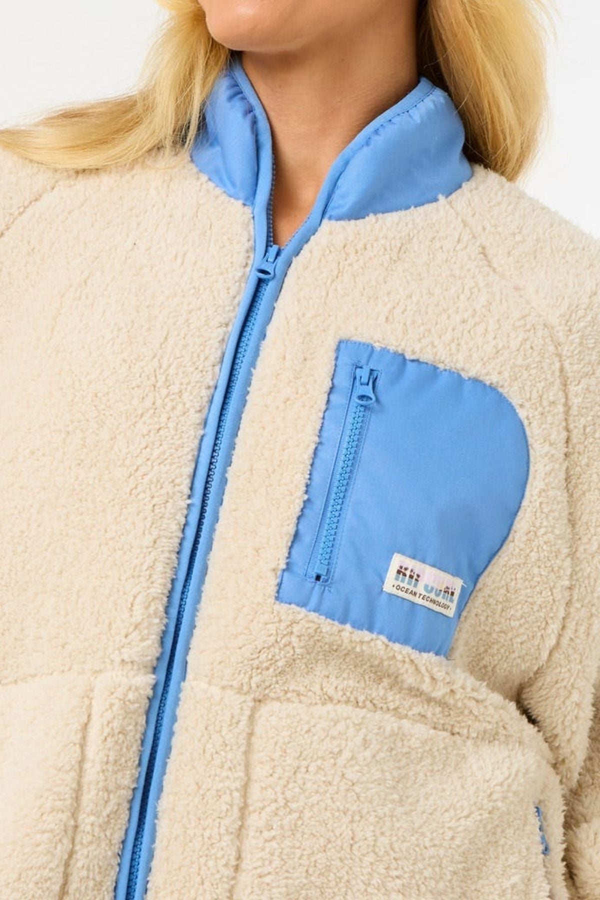 A person wears the Rip Curl Shore Break Polar Fleece, a cream 100% Polyester 300GSM relaxed fit pullover with bright blue zipper, collar, and chest pocket. Only their lower face and upper torso are visible.