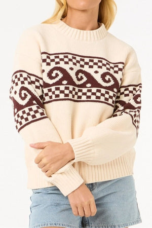 Wearing the Rip Curl Soleil Knit Crew, an oversized cream long sleeve with brown geometric and wave designs, paired with light blue denim shorts. The person's face is not shown.