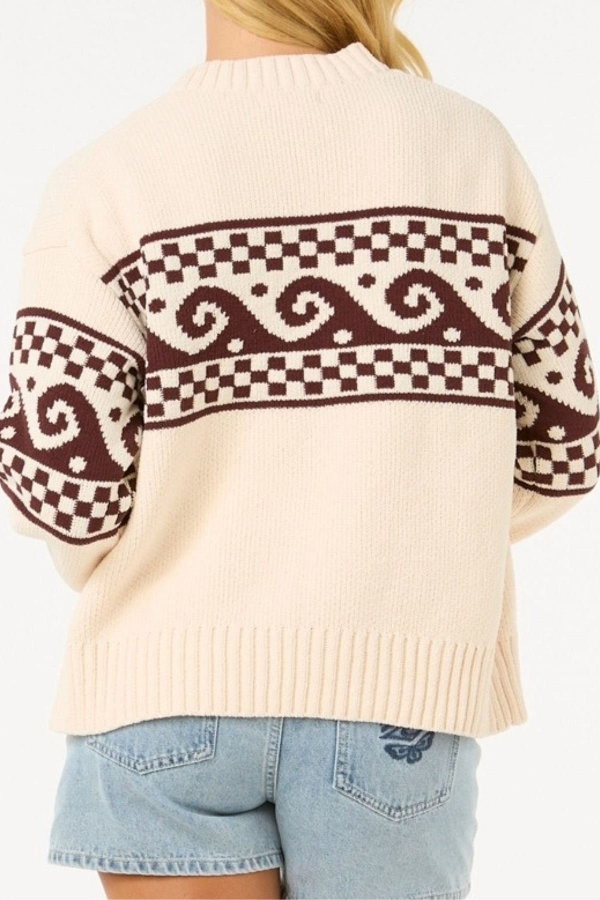 A person is seen from the back wearing the Rip Curl Soleil Knit Crew, an oversized cream long sleeve knit with brown geometric and wave designs on the upper back and sleeves, paired with light blue denim shorts.