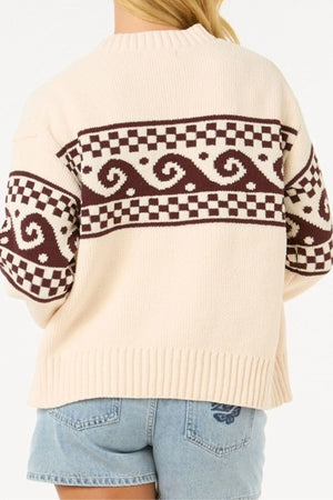 A person is seen from the back wearing the Rip Curl Soleil Knit Crew, an oversized cream long sleeve knit with brown geometric and wave designs on the upper back and sleeves, paired with light blue denim shorts.